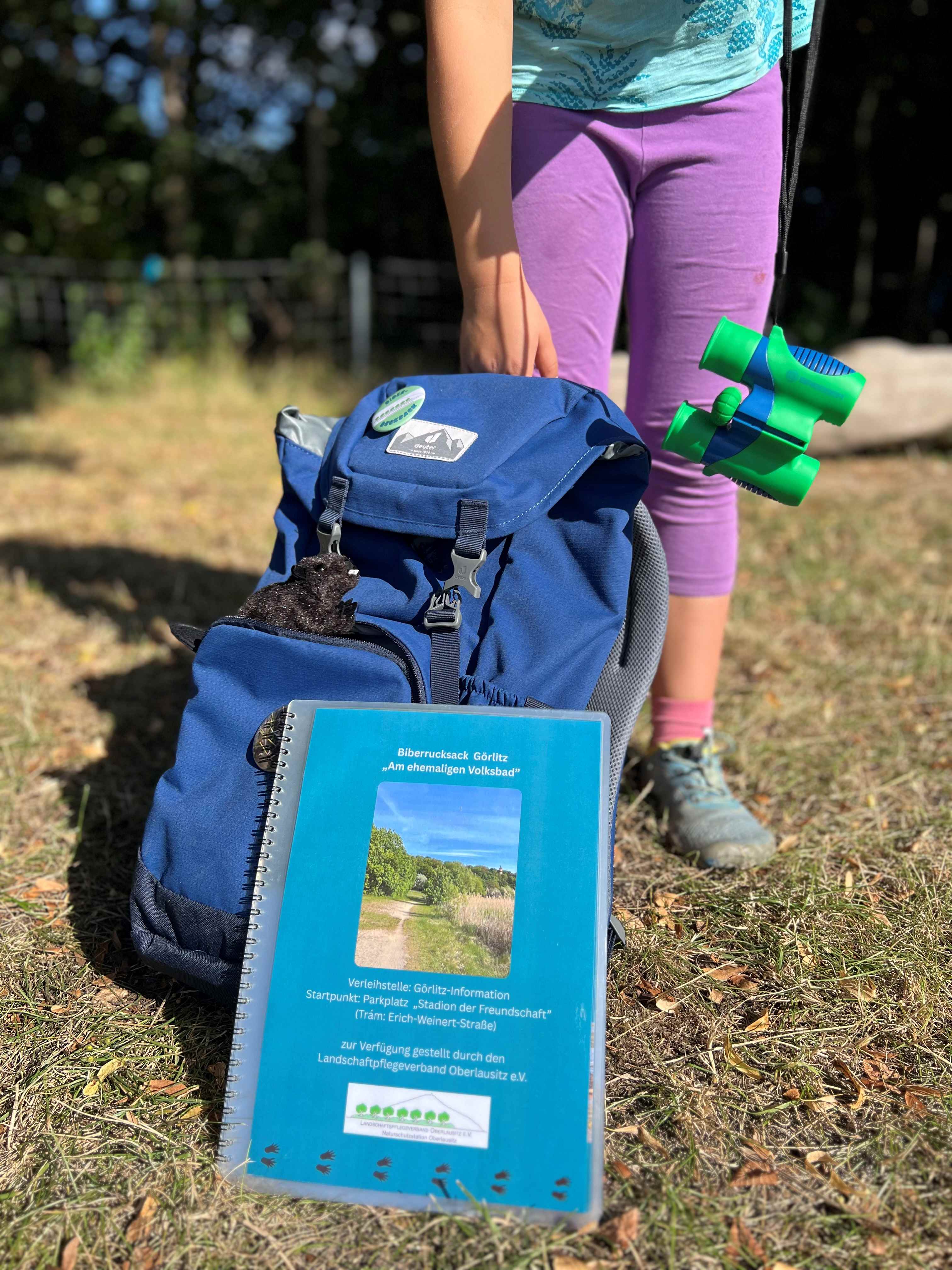 Eine Mappe und ein Rucksack stehen im Gras. Im Hintergrund ist ein Kind zu sehen